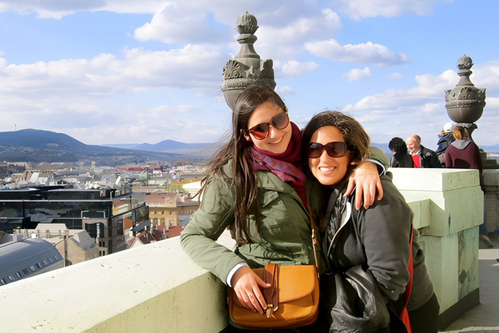 Two people with their arms around each other pose for the camera against a foreign city backdrop.