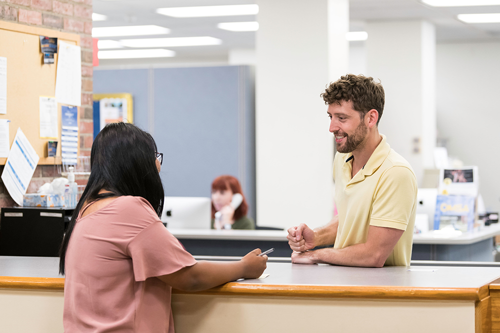 A person standing at a desk speaking to another person.